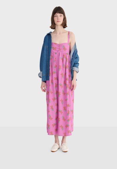 Jeune femme portant une robe midi rose à fleurs avec des bretelles spaghetti, des baskets blanches et une veste en jean posée sur ses épaules.