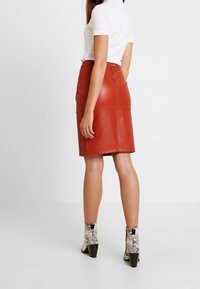 Červená kožená tužková sukně se zadním zipem, hladkou texturou a úzkým střihem. Skvěle ladí s kotníkovými botami se vzorem hadí kůže a bílým žebrovaným topem.