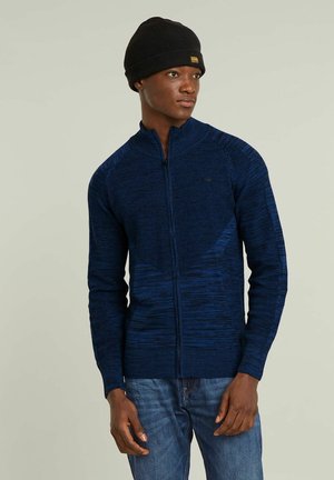 Pull en tricot zippé bleu marine avec un motif texturé, coupe ajustée et poignets côtelés. Porté avec un bonnet noir.