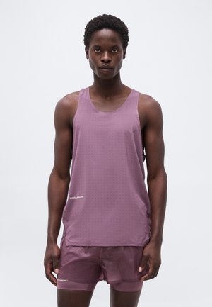 Αθλητικό άτομο που φοράει μοβ χωρίς μανίκια διάτρητη φανέλα και αντιστοιχία ελαφριά σορτς, στέκεται μπροστά σε απλό φόντο.