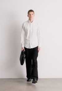 Λευκό πουκάμισο με κουμπιά και μαύρο παντελόνι, κρατώντας μαύρη τσάντα. Το πάτωμα είναι ανοιχτό γκρι, το φόντο άσπρο. Απλή, μοντέρνα ενδυμασία με λειτουργικό σχεδιασμό.