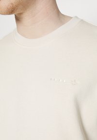 Sudadera de cuello redondo en color crema claro, con una textura suave, un sutil logotipo bordado y cinco pequeños puntos en fila en el pecho.
