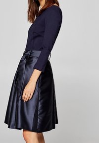 Kvinna som bär en marinblå långärmad topp och en blanka marinblå knälång kjol med ett knytbälte, stående i profil mot en grå bakgrund.