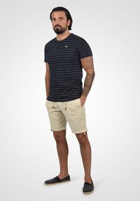 Majica s mornarskim prugama i kratkim rukavima, uparen s bež šorcem s pojasom na vezanje i bočnim džepovima, nošeno s tamnim slip-on cipelama.
