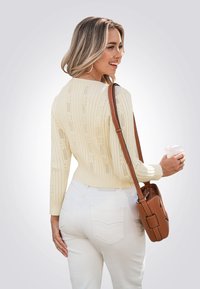 Crèmekleurige kabelgebreide trui met een open ruitontwerp, high-waist witte jeans en een bruine schoudertas. Iemand houdt een drankje in één hand.
