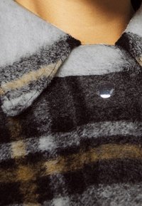 Camicia in misto lana con un motivo a quadri nei toni del grigio scuro, nero e marrone. Presenta un colletto a punta e una chiusura con bottoni trasparenti. Tessuto morbido e testurizzato.