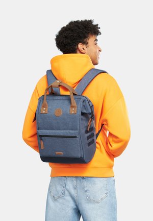 Zaino blu con dettagli in pelle marrone, un'unica maniglia superiore, tasca frontale con zip e spallacci imbottiti, indossato sopra un hoodie arancione.