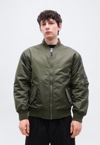 Νέος άνδρας που φοράει πράσινο μπουφάν bomber με φερμουάρ, ριμπ μανσέτες και γιακά, στέκεται μπροστά σε ένα απλό φωτεινό φόντο.