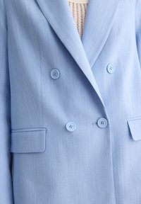 Blazer doppio petto azzurro chiaro, con due tasche frontali, bottoni blu e un tessuto a trama fine. Design sartoriale con rever affilati.
