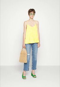 Pessoa vestindo um top amarelo sem mangas, jeans rasgados em azul claro, sapatos de salto abertos verdes, óculos de sol grandes verdes e segurando uma mochila de palha bege.