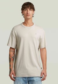 Beige t-shirt med kort ärm i mjukt tyg, med rund halsringning och en liten logotyp på vänster bröst. Avslappnad passform.