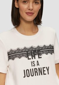 Witte katoenen t-shirt met een zwart kanten ontwerp over de borst, met daaronder in vette tekst "HET LEVEN IS EEN REIS".