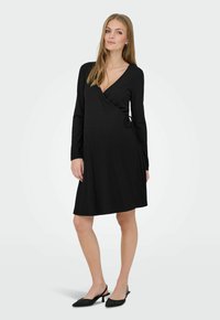 Femme portant une robe portefeuille noire à manches longues et des chaussures slingback noires à petit talon, debout devant un fond blanc uni.