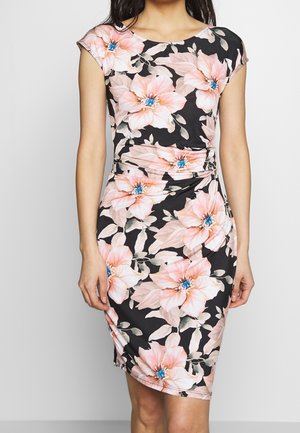 Kobieta w czarnej dopasowanej sukience z dużymi różowymi kwiatami z niebieskimi środkami, z krótkimi rękawkami i talią podkreśloną pasem.