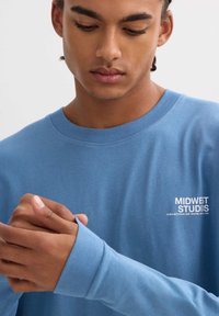 Camisa azul de manga larga hecha de algodón, con un cuello redondo y un logotipo en el pecho en blanco. La tela parece suave y lisa.