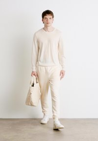 Beige gebreide trui en bijpassende joggingsbroek van zacht materiaal, gecombineerd met een lichtbeige totebag en witte sneakers, tegen een effen achtergrond.