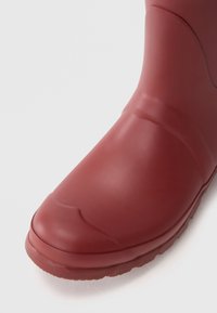 Botte en caoutchouc rouge avec une finition lisse et mate, un bout arrondi et des détails de couture subtils sur la partie supérieure et la semelle.