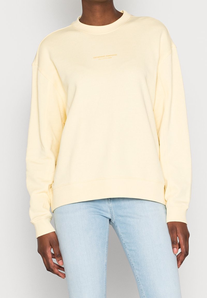 Personne portant un sweat-shirt à manches longues jaune pâle et un jean bleu clair, debout devant un fond clair uni.