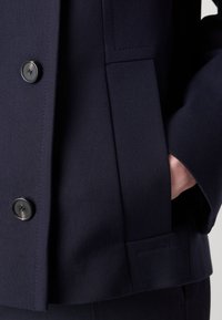 Blazer navy en tissu texturé, avec de grands boutons noirs et une poche latérale. Lignes épurées avec une coutume structurée évidente.