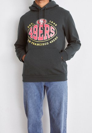 Person trägt dunkelgrauen San Francisco 49ers Hoodie mit rotem und gelbem Logo und blauen Jeans, Hände in der Kapuzen-Tasche.