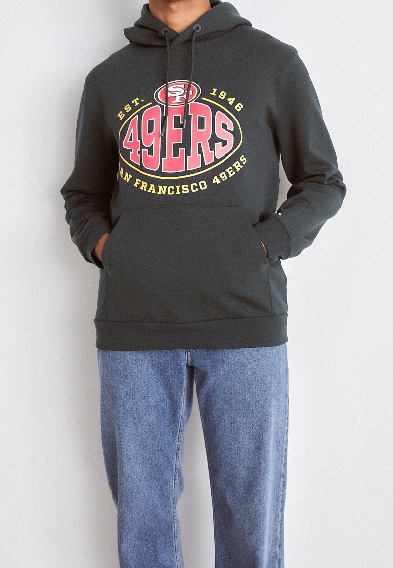 Personne portant un sweat à capuche gris foncé San Francisco 49ers avec un logo rouge et jaune et un jean bleu, mains dans la poche du sweat à capuche.