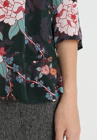 Blommig blus med korta ärmar; mörkgrön bas med rosa, röda och teal blommor; lättviktsmaterial; sidofickdetalj; i kombination med grå strukturerade byxor.