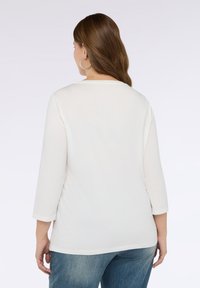 Kobieta z brązowymi włosami, nosząca srebrne kolczyki w kształcie kół, białą bluzkę z długim rękawem i niebieskie dżinsy, stojąca tyłem na jednolitym tle.