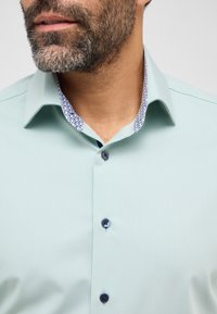 Mintgrünes Hemd mit texturierter Oberfläche, kontrastierendem gemustertem Innenkragen und marineblauen Knöpfen. Verfügt über einen klassischen Kragen und eine Knopfleiste auf der Vorderseite.