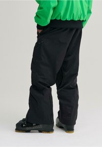 Pantaloni da sci neri con cerniere laterali e vita regolabile. Indossati con scarponi da sci neri con suole gialle. Il modello indossa una giacca verde.