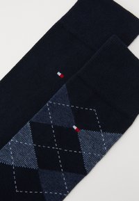 Marineblaue Socken mit Argyle-Muster in verschiedenen dunklen Blautönen, versehen mit einem kleinen rot-weiß-blauen Logo nahe der Oberseite. Weiche Strick-Texture.