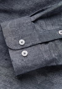 Poignet de chemise gris avec deux boutons blancs et motifs floraux sombres et feuilles sur un tissu texturé.