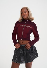 Kobieta o blond włosach nosi krótki burgundowy zamek i czarną mini spódniczkę w wzory, pozując z rękami na talii spódnicy.