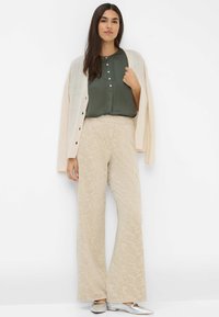Beige, gemusterte weit geschnittene Hose mit elastischem Bund, kombiniert mit einer dunkelgrünen Bluse und einem hellbeigen Cardigan. Silberne Schuhe setzen Akzente.