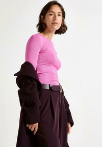 Top aderente a maniche lunghe di colore rosa acceso, indossato sotto un cappotto scuro. La modella porta pantaloni marroni larghi con una cintura nera larga e fibbia argentata.