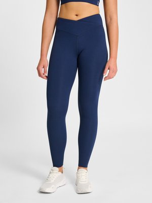 Vrouw draagt marineblauwe legging met hoge taille en witte sportschoenen, staand tegen een effen lichte achtergrond.