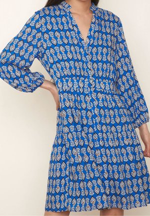 Kobieta ubrana w niebieską sukienkę do kolan z białymi i brązowymi wzorami liści, z długimi rękawami, dekoltem w serek i elastyczną talią, stojąca z jedną ręką na biodrze.