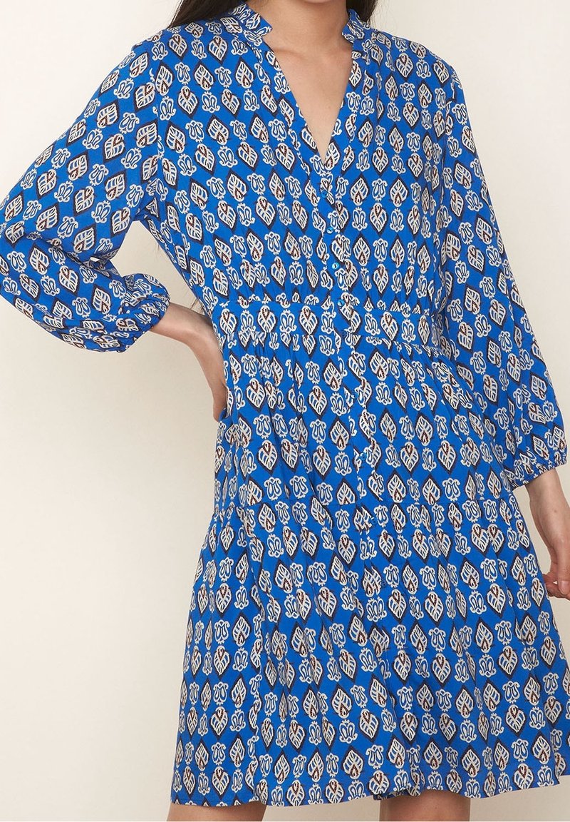 Femme portant une robe bleue mi-longue avec des motifs de feuilles blanches et marron, manches longues, col en V et taille élastique, debout avec une main sur la hanche.
