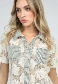 Chemisier en dentelle avec motifs floraux, accents tissés rayés sur le col et les poches, fermeture à boutons et manches courtes. Couleurs crème et noir.