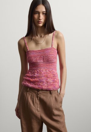 Jeune femme aux cheveux foncés et raides portant un caraco en maille rose texturée et un pantalon marron ample, debout devant un fond clair uni.