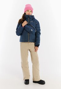 Blauwe spijkerjack met een capuchon, beige broeken, roze gebreide beanie en zwarte schoenen. De jas heeft een ritssluiting aan de voorkant en zichtbare stiksels.