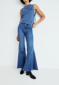Denim set koji se sastoji od topa bez rukava s bočnim vezicama i širokim trapericama. Oboje je u srednje plavoj nijansi s glatkom teksturom i suptilnim šavovima.