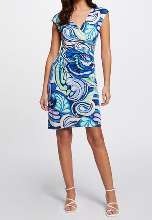 Kobieta w sukience kopertowej do kolan, bez rękawów, w niebieskie, zielone i fioletowe abstrakcyjne wzory w spirale, zestawione z białymi sandałami na obcasie.