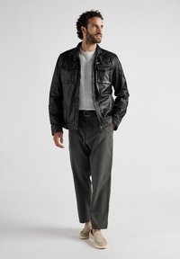 Schwarze Lederjacke mit zwei Brusttaschen, grauer Strickpullover und olivgrüne Hose. Das Modell posiert in einer entspannten Haltung und trägt leichte Schuhe.