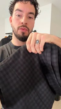 Crni karirani materijal drži se u ruci, s glatkom teksturom i blagim sjajem. Na ruci je prsten u obliku kruga s detaljnim dizajnom.