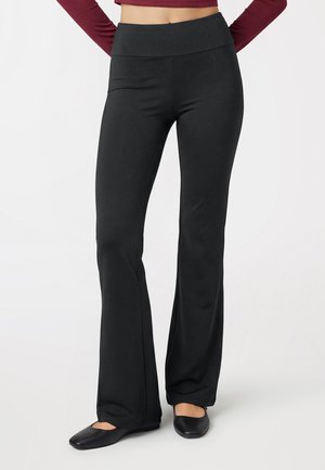 Osoba ubrana w czarne, wysokie spodnie z rozszerzanymi nogawkami, do tego czarne baleriny i długi, bordowy top odsłaniający brzuch, stoi z rękami na biodrach.