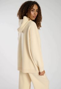 Sudadera beige de gran tamaño con una textura suave, que cuenta con capucha y mangas anchas, combinada con pantalones beige a juego, sobre un fondo neutro.