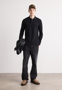 Μαύρο μακρυμάνικο πόλο μπλουζάκι, γκρι τζιν παντελόνι, καφέ αναπαυτικά παπούτσια και καπέλο καφέ. Το μοντέλο κρατάει μια μαύρη δερμάτινη ζακέτα. Ουδέτερο φόντο.