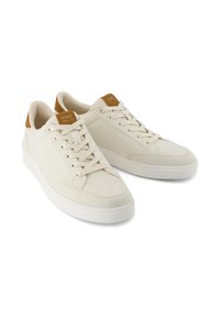 Beiga canvas sneakers med en vit gummisula, med en tan läderdetalj på hälen och snörning. TOMS-märket på tungan.