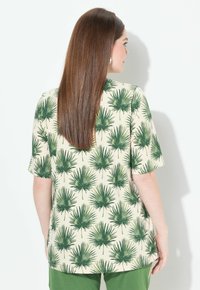 Top de manga corta con un fondo crema y grandes estampados de hojas de palma verdes. La tela parece suave; el diseño tiene un ajuste relajado.