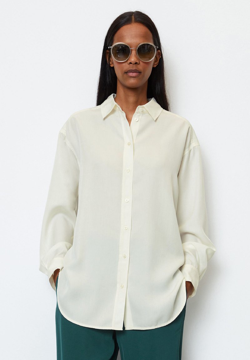 Light ivory shirt made of smooth fabric, featuring a button front, collar, and long sleeves. Model wears oversized sunglasses and green trousers.